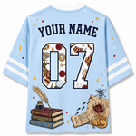 A light blue short-sleeve t-shirt shown from the back, featuring customizable text 'YOUR NAME' and the number '07' filled with wizarding-themed illustrations. The design includes a feather quill, books, a map, a cat, and a butterfly on a clean white background.