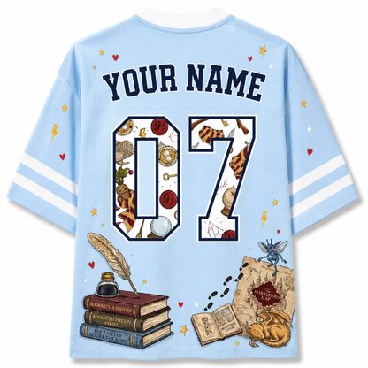 A light blue short-sleeve t-shirt shown from the back, featuring customizable text 'YOUR NAME' and the number '07' filled with wizarding-themed illustrations. The design includes a feather quill, books, a map, a cat, and a butterfly on a clean white background.