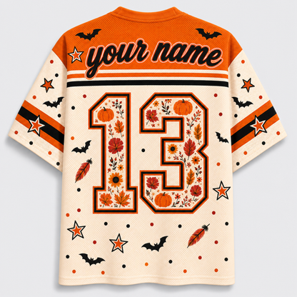 A short-sleeve mesh jersey-style shirt shown from the back on a light gray background. The shirt is cream with orange and black graphics, including the text 'your name', a large number 13 filled with autumn leaves and pumpkins, stars, bats, and feather motifs.