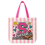 A pink and white striped canvas tote bag with bright pink handles, featuring a personalized graphic with a glittery pink letter 'S', the name 'Sarah', stars, hearts, a smiley face, books, and an apple on a white background.
