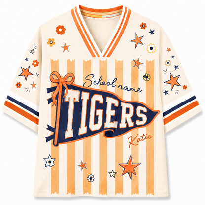A short-sleeve V-neck t-shirt with a cream base, orange and navy stripes, and a large 'TIGERS' banner graphic on the front. The shirt features decorative stars, flowers, and the text 'School name' and 'Katie' on the sleeves and body.