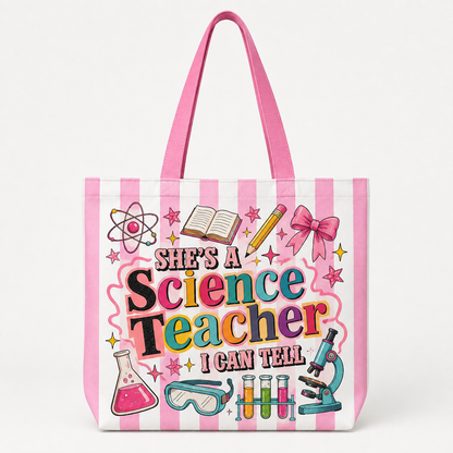 A pink and white striped tote bag featuring a colorful graphic that reads 'She's a Science Teacher I Can Tell' surrounded by science-themed illustrations including a microscope, beaker, test tubes, and an atom symbol, shown on a clean white background.