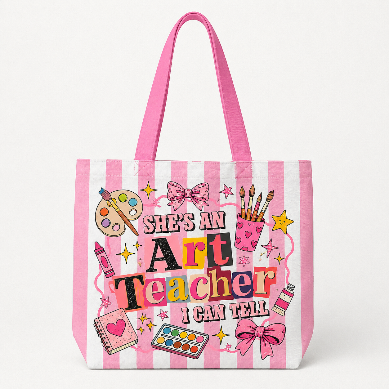 A pink and white striped canvas tote bag featuring a colorful graphic that reads 'SHE'S AN Art Teacher I CAN TELL' surrounded by art-themed illustrations like paintbrushes, a palette, crayons, and bows, shown on a clean white background.