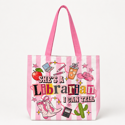 A pink and white striped tote bag with long pink handles, featuring a colorful graphic that reads 'SHE'S A Librarian I CAN TELL' surrounded by librarian-themed illustrations including books, an apple, a cowboy hat, and a cactus.