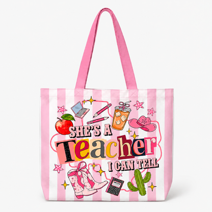 A pink and white striped tote bag with a colorful graphic that reads 'She's a Teacher I Can Tell' surrounded by school and western-themed illustrations, shown on a clean white background.