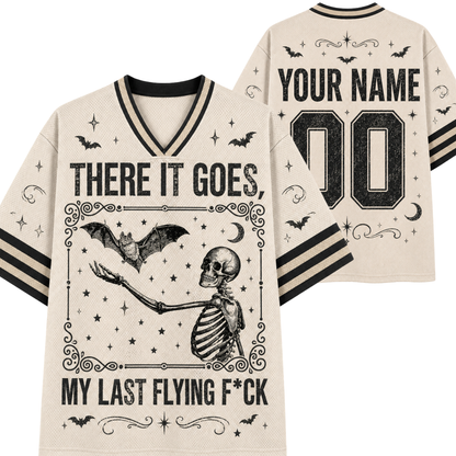 A cream-colored short-sleeve V-neck t-shirt with black graphic prints of a skeleton, bats, and stars, featuring customizable text on the front and back. The front reads 'THERE IT GOES MY LAST FLYING F*CK' with a central skeleton graphic, and the back shows 'YOUR NAME' and '00'.