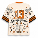 A cream-colored short-sleeve t-shirt with orange and black accents, featuring a Halloween-themed graphic on the back. The design includes the text '13 Spooky Season' in orange and black, a cartoon bull in a white cape, spiders, bats, stars, and flowers.