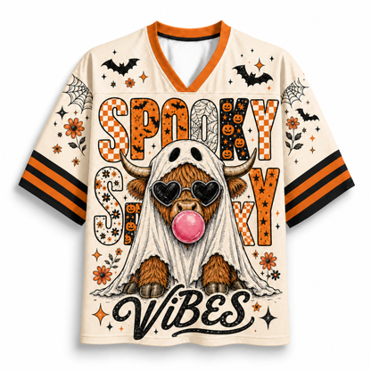 A short-sleeve V-neck jersey-style shirt with a Halloween graphic print. The cream-colored shirt features orange and black striped sleeves, a white ghost graphic, bats, spiders, and stars, and the text 'Spooky Sexy Vibes' with a cartoon highland cow wearing heart-shaped sunglasses and a pink bubblegum bubble.