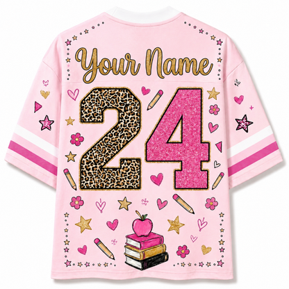 A pink short-sleeve t-shirt shown from the back, featuring customizable text 'Your Name' in gold glitter, the number '24' with leopard print and pink glitter fill, and school-themed illustrations including hearts, stars, pencils, books, and an apple.