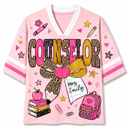 A pink short-sleeve V-neck t-shirt with a white collar, featuring a colorful graphic print on the front. The design includes the word 'COUNSELOR' in leopard-print and pink letters, a large leopard-print bow with a pink apple, a pencil, a notebook that says 'Mrs.