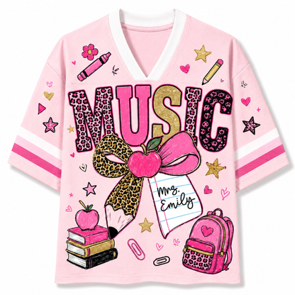 A pink V-neck t-shirt with a school-themed 'MUSIC' graphic, featuring leopard print lettering, a large pink apple bow, a notebook reading 'Mrs. Emily', and school doodles like stars, pencils, and a backpack on a white background.