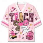 A pink V-neck t-shirt with a school-themed graphic print featuring the word 'TEACH' in leopard print and gold glitter textures, a pink bow, an apple, books, a backpack, and a notebook that reads 'Mrs. Emily'.