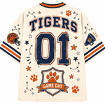 A cream-colored short-sleeve sports jersey shown from the back, featuring 'TIGERS' and '01' in navy blue with orange trim, along with orange paw prints, stars, a football helmet, a megaphone, and a 'GAME DAY' crest.