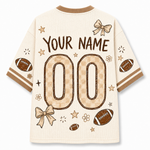 A cream-colored mesh short-sleeve jersey shown from the back, featuring customizable text 'YOUR NAME' and large '00' placeholders in a tan checkered pattern. The design includes brown footballs, beige bows, stars, and stripes on the sleeves.
