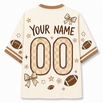 A cream-colored mesh short-sleeve jersey shown from the back, featuring customizable text 'YOUR NAME' and large '00' placeholders in a tan checkered pattern. The design includes brown footballs, beige bows, stars, and stripes on the sleeves.