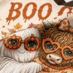 Close-up of a cozy autumn-themed textile design featuring a white ghost and a fluffy brown animal wearing heart-shaped orange glasses, with the word "BOO" in orange dripping letters on a cream background.