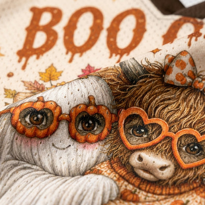 Close-up of a cozy autumn-themed textile design featuring a white ghost and a fluffy brown animal wearing heart-shaped orange glasses, with the word "BOO" in orange dripping letters on a cream background.