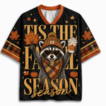 A black short-sleeve V-neck t-shirt with orange trim, featuring a festive autumn graphic of a raccoon in a beanie and scarf holding a pumpkin and a hot drink, surrounded by maple leaves, pumpkins, and the text 'TIS THE FALL SEASON'.