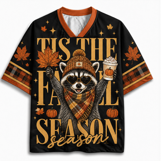 A black short-sleeve V-neck t-shirt with orange trim, featuring a festive autumn graphic of a raccoon in a beanie and scarf holding a pumpkin and a hot drink, surrounded by maple leaves, pumpkins, and the text 'TIS THE FALL SEASON'.