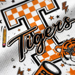 Close-up of a white athletic jersey or fabric featuring a large orange and black checkered letter T, the word 'Tigers' in black script, stars, lightning bolts, a pencil, and part of a tiger illustration.