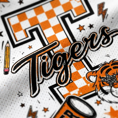 Close-up of a white athletic jersey or fabric featuring a large orange and black checkered letter T, the word 'Tigers' in black script, stars, lightning bolts, a pencil, and part of a tiger illustration.
