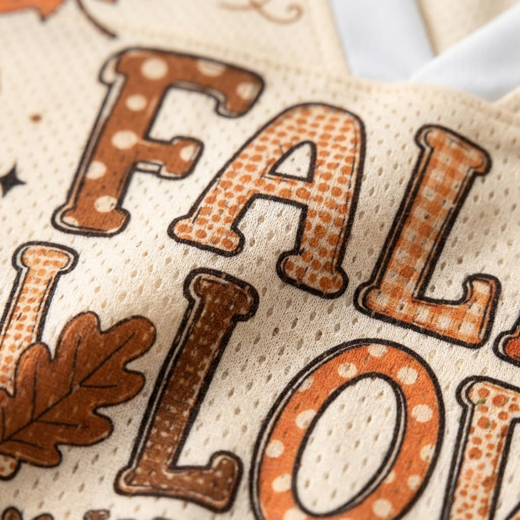 Close-up of a cream-colored mesh fabric with a 'FALL' word design in brown and orange, featuring polka dots, gingham, and solid textures, alongside a brown leaf motif.