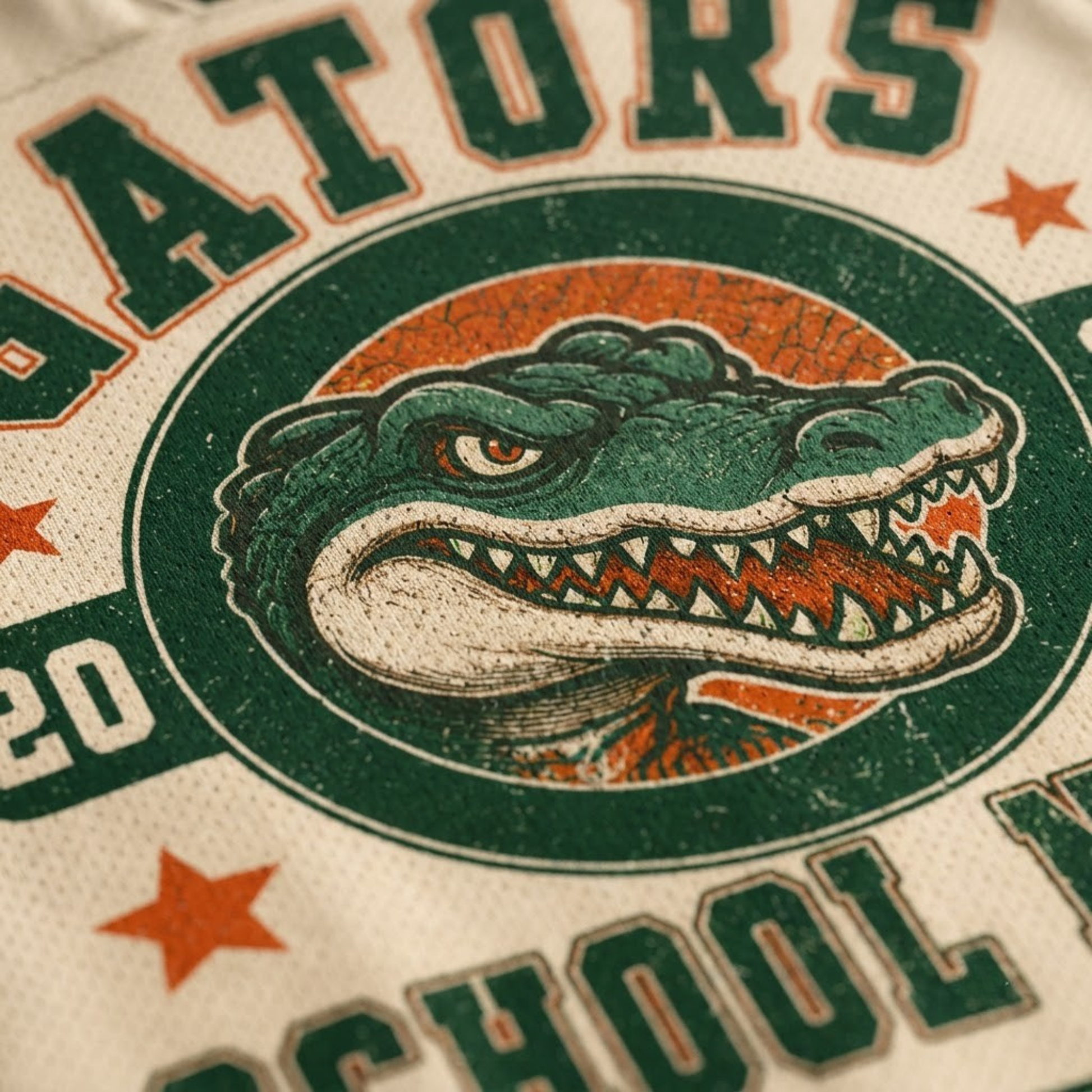 Close-up of a distressed vintage-style graphic print featuring an alligator mascot inside a circular emblem, with green lettering and orange stars on a cream fabric background.