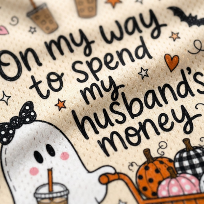 Close-up of a beige knit fabric with a printed Halloween-themed quote that reads 'On my way to spend my husband's money.' The design includes a cartoon ghost holding a drink, pumpkins, stars, bats, and small decorative motifs.