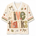 A short-sleeve V-neck t-shirt with a cream-colored base, featuring a 'give thanks' graphic in autumn-themed colors (coral, leopard print, olive green, beige) and decorated with fall motifs like acorns, leaves, and flowers on the sleeves and chest.