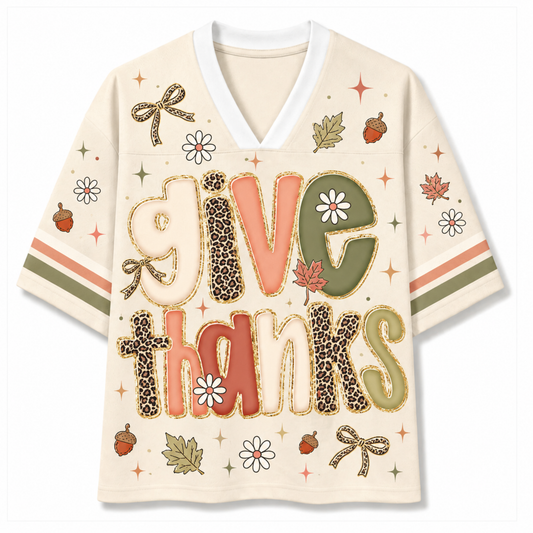 A short-sleeve V-neck t-shirt with a cream-colored base, featuring a 'give thanks' graphic in autumn-themed colors (coral, leopard print, olive green, beige) and decorated with fall motifs like acorns, leaves, and flowers on the sleeves and chest.