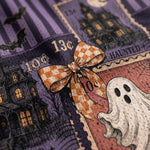 Close-up of a Halloween-themed fabric print featuring vintage postage stamps, a purple haunted house, a white ghost, bats, and an orange-and-white checkered bow.