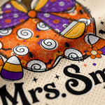 Close-up of a printed fabric design featuring a large purple and orange bow, candy corn, spiderwebs, black spiders, and the partial text 'Mrs. Sm' on a cream mesh background.