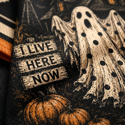 Close-up of a Halloween-themed fabric print featuring ghost figures, pumpkins, a haunted house, and a wooden sign that reads 'I LIVE HERE NOW' in black text.