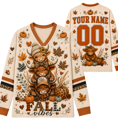 A long-sleeve V-neck sweater shown from both front and back on a white background. The cream base has autumn-themed graphics including stacked Highland cows, pumpkins, leaves, acorns, and the words 'FALL vibes' and 'YOUR NAME 00'.