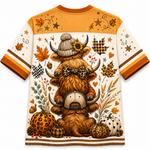 A short-sleeve crewneck t-shirt featuring a detailed autumn-themed graphic print of a shaggy Highland cow wearing a knit hat and leopard-print bow tie, surrounded by pumpkins, leaves, stars, and plaid patterns on an off-white background.