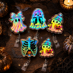 Five holographic Halloween-themed badge reels arranged on a dark wooden surface with spooky props. The designs include a ghost with a bow, a rainbow ghost, a ghost holding a pumpkin and beanie, a rib cage with a black heart, and a ghost in a striped beanie holding a pumpkin.