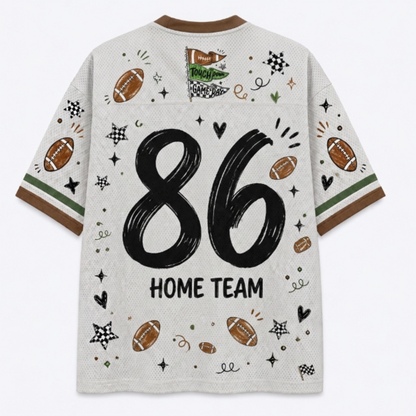 A white mesh short-sleeve t-shirt with a brown crew neck and sleeve cuffs, featuring a large black '86' and 'HOME TEAM' text on the back, decorated with football illustrations and sports doodles.