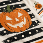 A close-up of a Halloween-themed fabric napkin or cloth featuring a large orange jack-o'-lantern with a green stem, black brushstroke stripes, white polka dots, and a small pumpkin motif in the corner.
