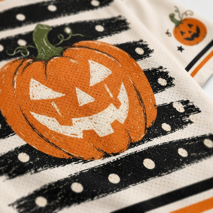 A close-up of a Halloween-themed fabric napkin or cloth featuring a large orange jack-o'-lantern with a green stem, black brushstroke stripes, white polka dots, and a small pumpkin motif in the corner.