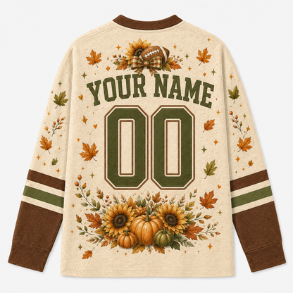A long-sleeve cream-colored sports jersey shown from the back, featuring customizable text 'YOUR NAME' and number '00' in dark green varsity lettering. The design is decorated with autumn-themed graphics including sunflowers, pumpkins, maple leaves, and a football motif on the shoulders, with green and brown stripes on the sleeves.