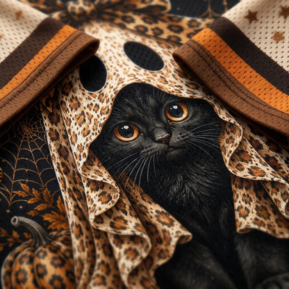 A black cat with large amber eyes peering through a leopard-print ruffled hood, styled over a dark patterned fabric with spiderwebs and pumpkins.