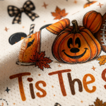 A close-up of a cream-colored fabric featuring a festive autumn pattern with a smiling jack-o'-lantern pumpkin, a slice of pumpkin pie, orange leaves, stars, and the text 'Tis The Season'.