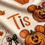 Close-up of a cream-colored garment with orange text reading 'Tis', orange pumpkin illustrations with Mickey Mouse-style ears, autumn leaves, and a black-and-white plaid bow.
