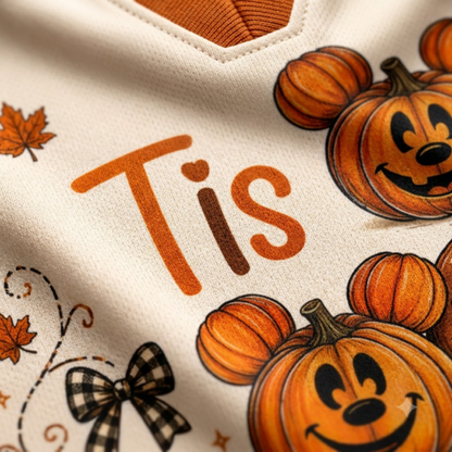 Close-up of a cream-colored garment with orange text reading 'Tis', orange pumpkin illustrations with Mickey Mouse-style ears, autumn leaves, and a black-and-white plaid bow.