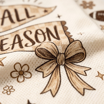 Close-up of a cream-colored fabric product featuring a brown printed design with the words 'ALL SEASON', stars, flowers, and a large bow motif.