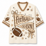 A short-sleeve V-neck athletic-style top in warm cream with tan trim and a football-themed graphic. The front features the words “FOOTBALL SEASON” over a tan banner, with illustrated footballs, bows, stars, and small flowers.