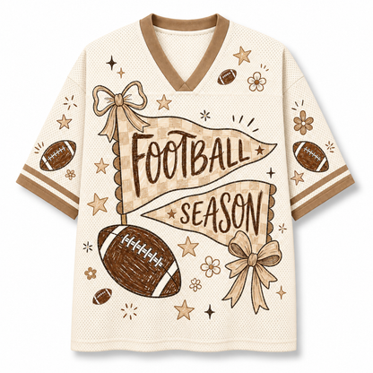 A short-sleeve V-neck athletic-style top in warm cream with tan trim and a football-themed graphic. The front features the words “FOOTBALL SEASON” over a tan banner, with illustrated footballs, bows, stars, and small flowers.
