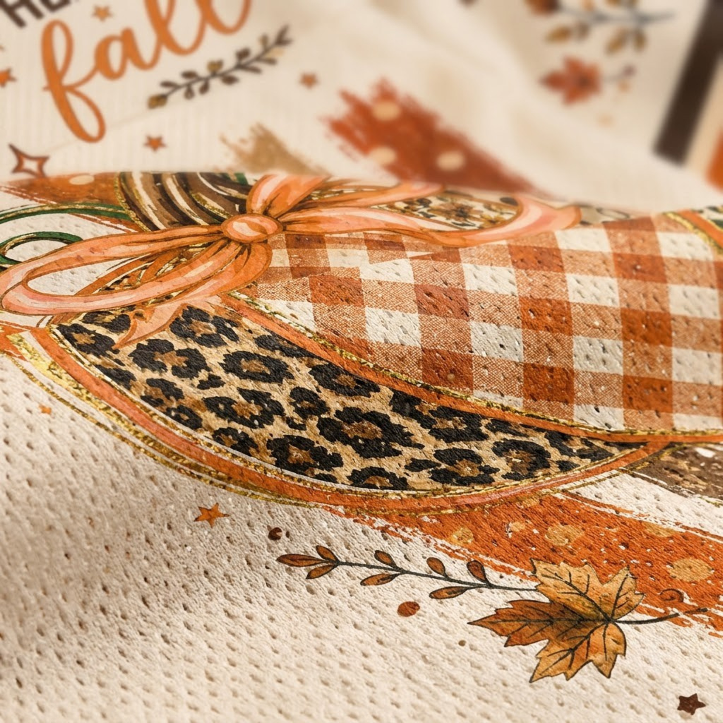 Close-up of a printed fabric or textile featuring an autumn-themed design with a gingham check pattern, leopard print, orange ribbon bow, leaves, stars, and partial text on a cream background.