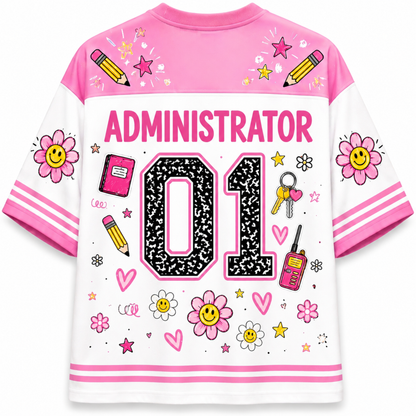 A short-sleeve athletic-style shirt shown from the back, featuring a white and pink base with cartoon graphics including flowers, pencils, stars, hearts, a walkie-talkie, keys, and the word 'ADMINISTRATOR' above large '01' lettering.