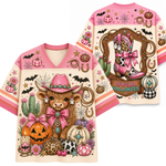 A pink and cream V-neck t-shirt with a detailed 'Cowgirl Halloween' graphic print, shown in both front and back views. The design features a highland cow in a pink cowboy hat, pumpkins, cacti, bats, and horseshoes.
